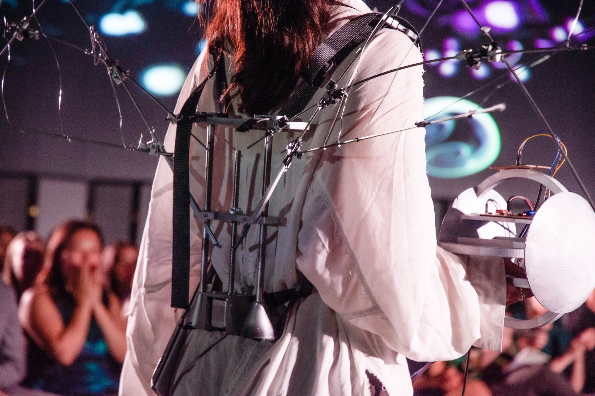 Close-up of a model wearing a mechanical, wing-like apparatus mounted on a harnessed frame over a white garment. The device extends from the back and includes structural rods, joints, and a spherical electronic component held in one hand.